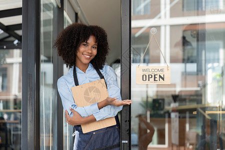 店铺开业展板小企业非洲女老板面带微笑 同时转向咖啡馆开业的标志 穿着围裙的快乐的美国黑人女服务员企业家在门上签名工作成人职业人士服务店铺咖啡背景