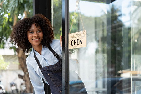 店铺开业展板小企业非洲女老板面带微笑 同时转向咖啡馆开业的标志 穿着围裙的快乐的美国黑人女服务员企业家在门上签名入口商业员工女性餐厅咖啡师女背景