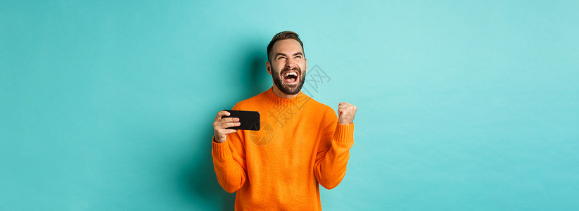 男人在手机上获胜 拳手泵和欢欣胜利 用满意的面孔持有智能手机 绿松石背景等形象喜悦标识电话潮人购物黑发工作室促销男性情感背景图片