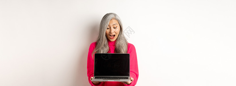 防癌宣传手册电子商务概念 以灰色头发震惊的亚洲祖母 在网上检查宣传手册 展示笔记本电脑黑屏 站在白背景上站立企业家互联网毛衣时尚快乐技术购物背景