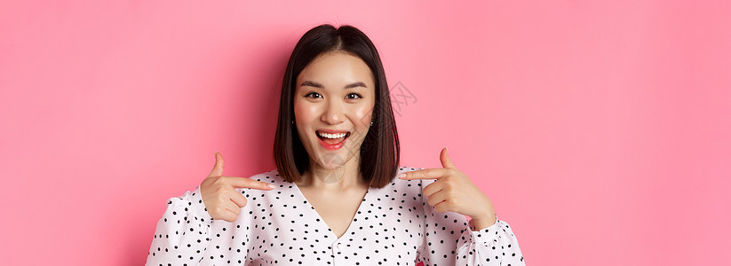 特写 快乐而自信的亚洲女性指着自己 微笑着自信 站在粉色背景上工作室冒充广告横幅情绪情感理发青少年购物皮肤背景图片