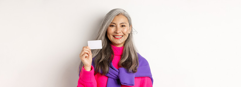 穿着时装 微笑和展示银行塑料信用卡 站在白色背景之上的漂亮亚洲高龄女士近身露脸 笑声女性乐趣母亲毛衣祖母信用奶奶时尚广告成人背景图片