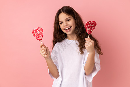 两颗棒棒糖满足的小女孩拿着两颗心形棒棒糖 用快乐的表情看着相机背景