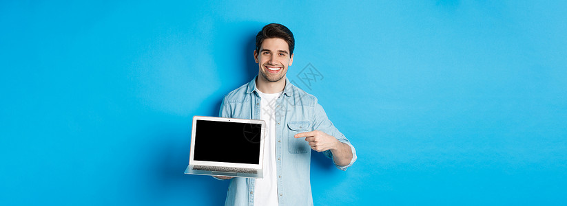 英俊的年轻人指着电脑屏幕 微笑着高兴 在互联网或网站上展示促销 站在蓝色背景上成人胡子工作室闲暇快乐技术情绪商业男性广告背景图片