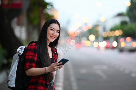 背包手机背着背包的年轻亚洲女人 用智能电话叫计程车 站在街上背景