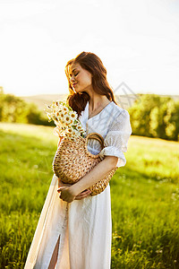 在一个田野日落的背景下 一个带满篮子雏菊的笑女站在一束光衣中背景图片