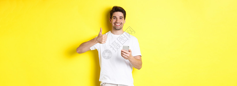 快乐满意的男人拿着智能手机 竖起大拇指表示赞同 在网上推荐一些东西 站在黄色背景上黑发互联网电话手势工具商业闲暇促销男性成人背景图片