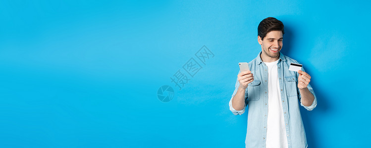帅气的快乐男人在网上买东西 拿着信用卡和手机 在网上购物 站在蓝色背景上胡子手势潮人胡须银行金融闲暇工作室促销成人背景图片