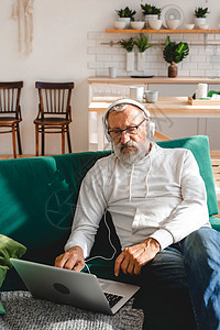穿着连帽衫和眼镜的英俊老男人正在使用笔记本电脑 并微笑地在家中听坐在沙发上的音乐头发房间成人会计老年网络经理退休工具互联网背景图片