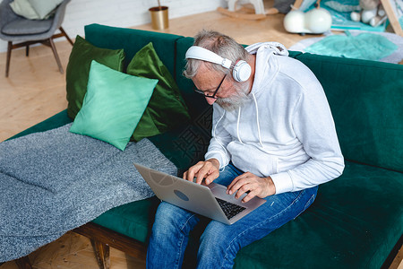穿着连帽衫和眼镜的英俊老男人正在使用笔记本电脑 并微笑地在家中听坐在沙发上的音乐职场企业家长椅会计男性经理头发互联网技术工具背景图片