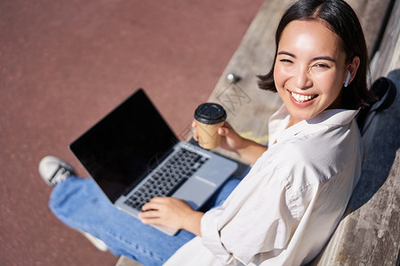 专享福利美丽的亚洲年轻女学生 带着笔记本电脑坐在公园的长椅上 喝外卖咖啡 听音乐 远程工作 在户外学习治疗微笑女性女孩自由数字学生福利享背景