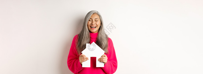 房地产 快乐的亚洲祖母闭着眼睛笑着 拿着纸屋模型 梦想着纸屋模型 站在白色背景上时尚老年母亲女性派对化妆品毛衣客户公寓奶奶背景图片