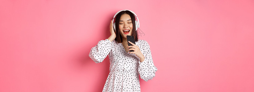可爱的亚洲女性玩卡拉 OK 应用程序 用手机唱歌 戴着耳机 穿着粉色背景的裙子皮肤音乐学生情感横幅工作室卡拉ok理发演员黑发背景图片