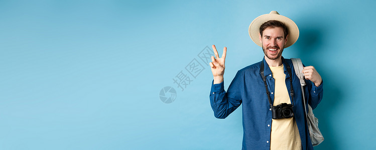 快乐的英俊男子在暑假时拍照 展示和平标志和微笑 戴草帽并持有蓝色背景旅游背包 AC 5   infol org帽子相机商业旅行成背景图片