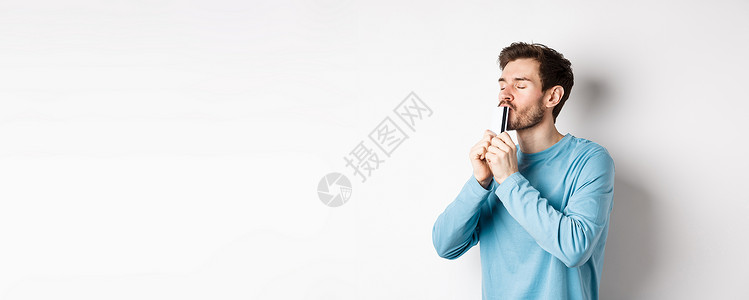 年轻男子亲吻塑料信用卡时面满脸满意 穿着蓝轻便衬衫站在白色背景上站立商业胡子银行工作室生活发型胡须成功情绪促销背景图片
