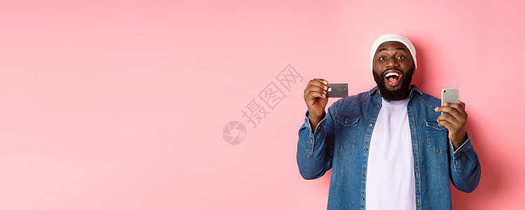 在网上购物 快乐的留胡子的非洲美国人微笑 展示信用卡和购买智能手机 站在粉红色背景面部站立着成人银行广告横幅电话促销胡须网络技术背景图片