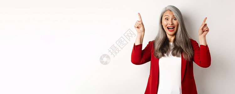 头发灰白 穿着红色西装外套 手指向上 惊讶地微笑 展示促销优惠 白色背景的成熟亚洲女商务人士退休祖母员工人士雇主套装工人经理工作背景图片
