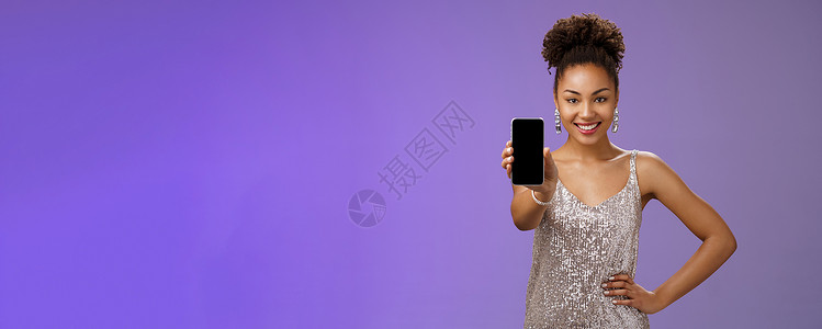 时髦优雅的非洲裔美国女性穿着闪闪发光的银色连衣裙 手握腰部自信的姿势 高兴地微笑着伸出手臂 展示智能手机显示屏 看看酷炫的应用程图片