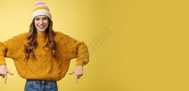 友好的乐于助人的漂亮的 20 多岁女大学生戴着帽子保暖毛衣微笑高兴地向您展示很棒的促销食指向下推荐底部广告温暖名声女性橙子新年大背景图片