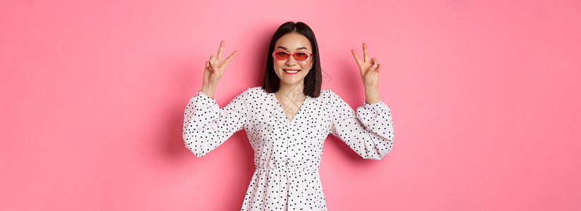 旅游和生活方式的概念 暑假中美丽的亚洲女性 展示和平标志 微笑快乐 戴着时髦的太阳镜 粉红色背景理发促销情绪横幅皮肤工作室冒充情背景图片