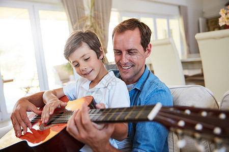 他吉他的父亲父亲和儿子的音乐乐趣 一位父亲和他年幼的儿子一起坐在家里的客厅里弹吉他背景