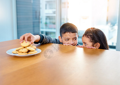 看看我得到了什么让我们享受 两个淘气的小孩在厨房桌子上偷饼干 在家里吃饼干背景图片