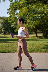 活跃的亚洲女孩 穿健身衣 在公园锻炼 穿着智能手机和水瓶运动服行走慢跑者日落跑步女性太阳赛跑者训练成人女士电话背景图片