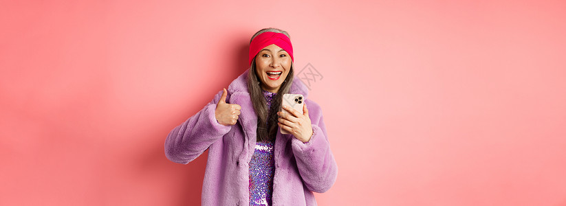 网上购物和时尚概念 快乐的亚洲祖母检查了惊人的宣传 用智能手机指着手指 看得惊讶 粉红色背景 笑声促销广告女性派对喜悦微笑头巾电背景图片