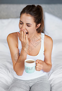 还这么困 一位年轻女子坐在床上喝着咖啡打个大哈欠背景图片