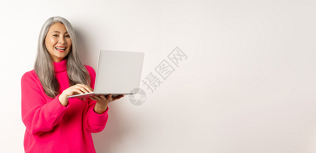 利用笔记本电脑工作 对着镜头笑笑和微笑 站在白人背景之上的美丽的亚洲女性高级创业者商务企业家老年祖母毛衣商业电子商务互联网促销网背景图片