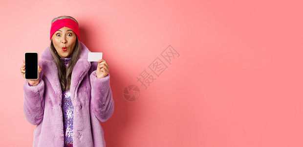 特别促销 兴奋的亚洲高级女性展示空白的智能手机屏幕和塑料信用卡 查看在线报价 粉红色背景电话祖母微笑银行购物技术广告奶奶头巾时尚背景图片