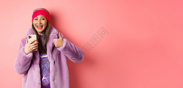 网上购物和时尚概念 快乐的亚洲祖母检查了惊人的宣传 用智能手机指着手指 看得惊讶 粉红色背景 笑声裙子技术喜悦女性标识乐趣化妆品背景图片