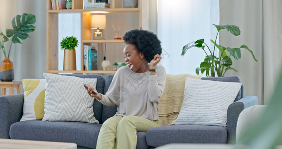 购物款欢非洲妇女坐在家里的沙发上庆祝一份新工作 一位年轻女性的贷款通过她手机上的电子邮件获得批准 一位快乐而兴奋的女士在沙发上为晋升而欢背景