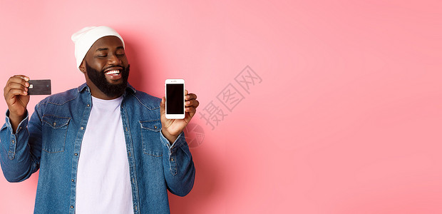 满意的黑人点头 微笑和看着电话 展示信用卡和智能手机屏幕 站在粉红背景上 掌声   un org互联网帽子情感金融男人银行广告学背景图片