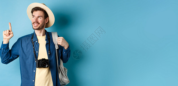 花艺相机徽标穿着暑假帽子 看和指着徽标 笑笑高兴 拿着相机和背包站在蓝底背景上Blue Brif胡须旅游男性男人成人旅行快乐成功乐趣生活背景