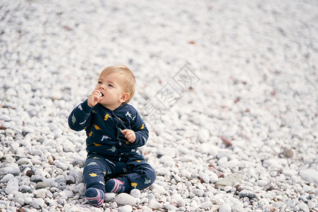 身穿蓝色工装裤的可爱婴儿坐在鹅卵石滩上 头向左转 手里拿着一块鹅卵石 将它拉进嘴里背景图片