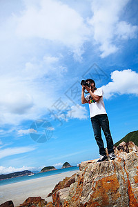 美化照片素材一个年轻男子的肖像 站在海滩上拿着相机全景乐趣摄影师照片岩石微笑石头季节观光丁字裤背景