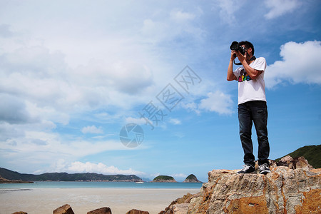 美化照片素材一个年轻男子的肖像 站在海滩上拿着相机棕褐色泳装季节石头身体游客观光乐趣男人岩石背景