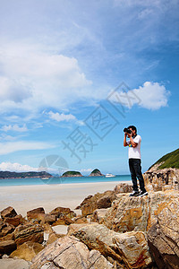美化照片素材一个年轻男子的肖像 站在海滩上拿着相机美化青少年身体季节岩石全景旅行石头棕褐色丁字裤背景