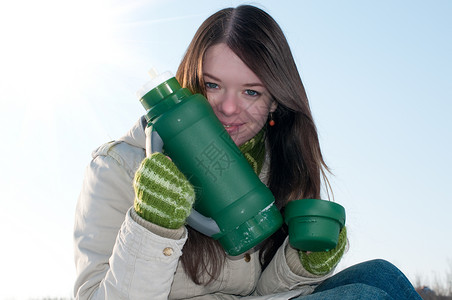 女孩在冬季野餐时 喝着热茶和热水太阳保温瓶天空蓝色外套青少年热水瓶女士女性长发背景图片
