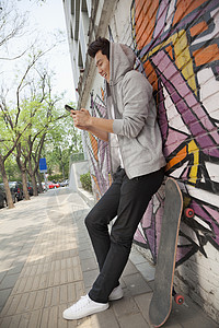 年轻人再次靠在满是涂鸦的墙上 用手机发短信 滑板就在他旁边背景图片