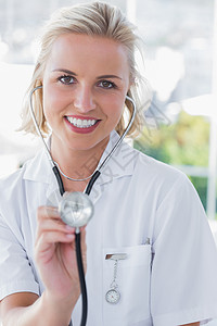 微笑的护士展示她的听诊器办公室金发医务室卫生快乐女性诊所浅色头发女士背景图片