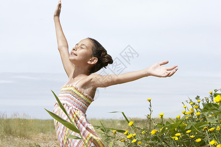 收腰裙子怀着手臂伸展的少女在田野中站在花朵旁 享受阳光光照耀背景