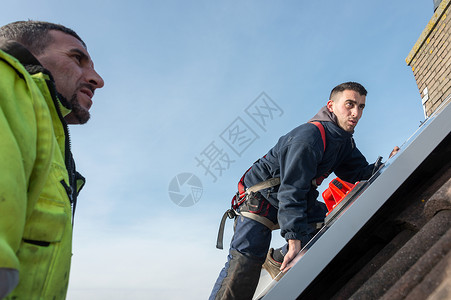 安全屋男人把太阳能电池板放在屋顶上控制板力量工人房子住宅梯子维修腰带工具蓝色背景