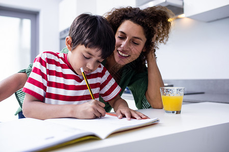 母亲帮助孩子做家庭作业休闲橙汁服装男性男生铅笔学习写作厨房微笑背景图片