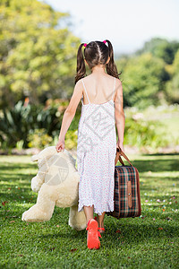 年轻女孩带着手提箱和泰迪熊在公园中行走背景图片