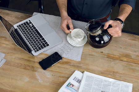 男人用笔记本电脑和报纸把咖啡倒在桌子上的杯子里的部分观点人士裁剪自由职业者商业远程手机工作者商务成人办公在家办公高清图片素材