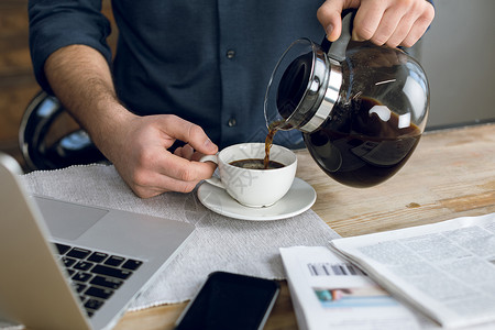 用笔记本电脑和智能手机在桌上用杯子倒咖啡的人的部分视图早晨高清图片素材