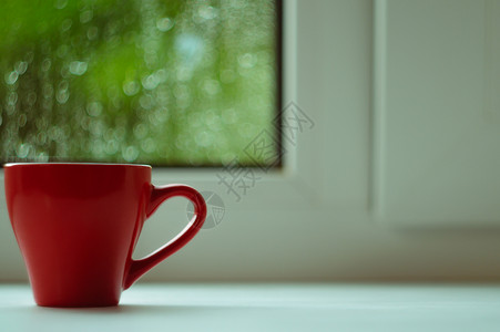 窗台边框白色窗台上的红咖啡杯 背景窗口和雨中 杯子位于边框左侧绿色红色太阳窗户咖啡中心背景