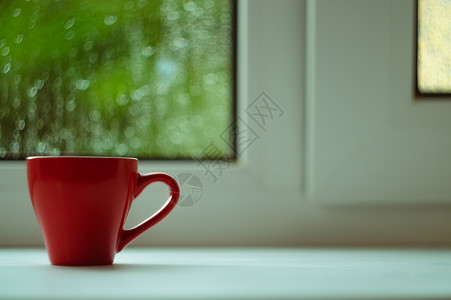 窗台边框白色窗台上的红咖啡杯 背景窗口和雨中 杯子位于边框左侧中心绿色咖啡窗户红色太阳背景